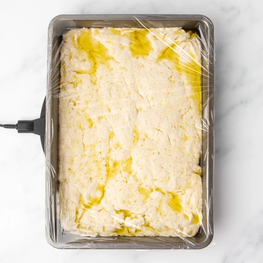 Pale dough shaped into a loose rectangle in baking pan with plastic wrap covering the top and pools of olive oil on top