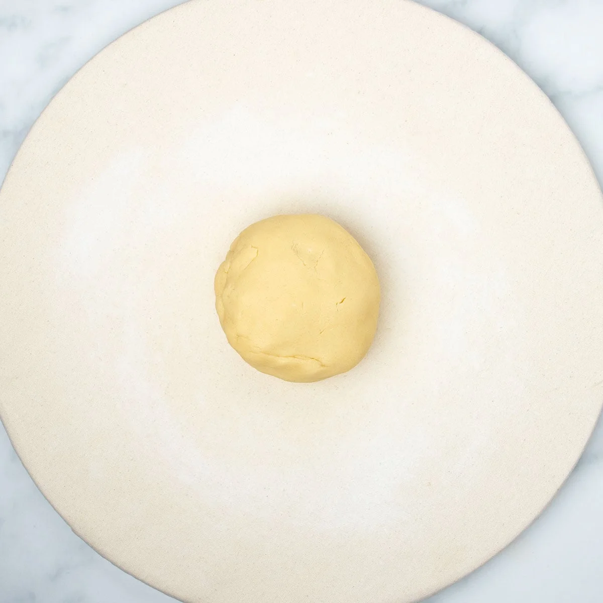 Pale yellow disk of raw sugar cookie dough in center of round canvas covered pastry board.
