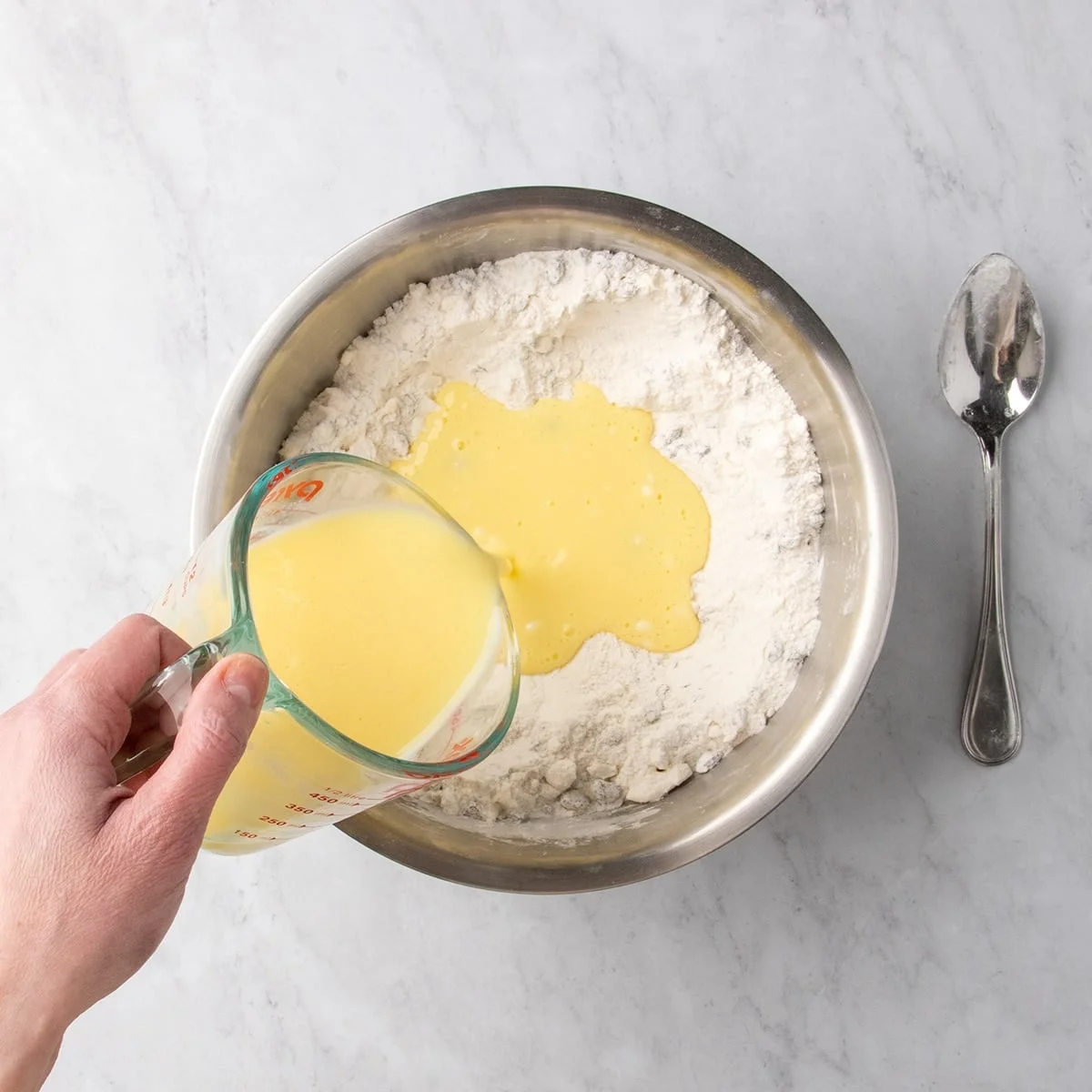 Pale yellow liquid being poured from a glass measuring cup into a bowl of mixed dry ingredients with grated butter and raisins.