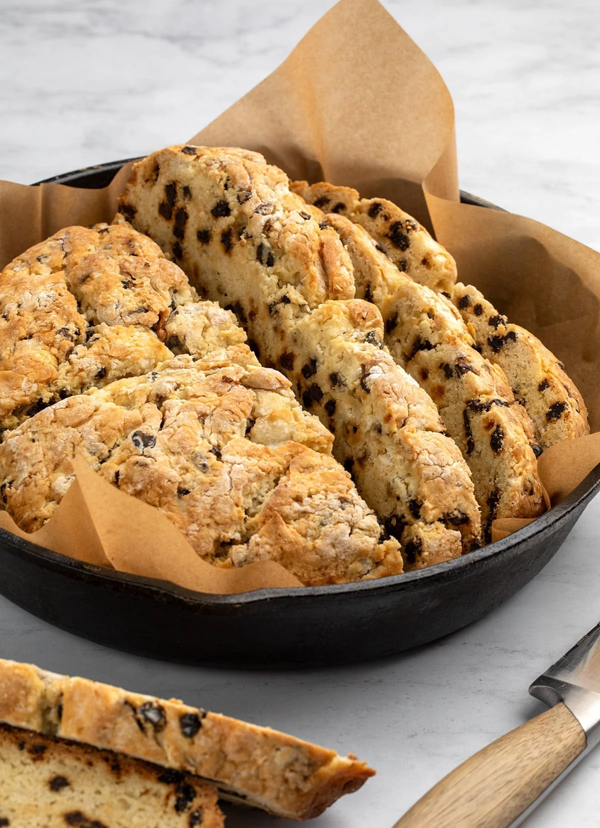 Partially sliced gluten free Irish soda bread in black cast iron skillet on marble surface with bread knife with brown handle