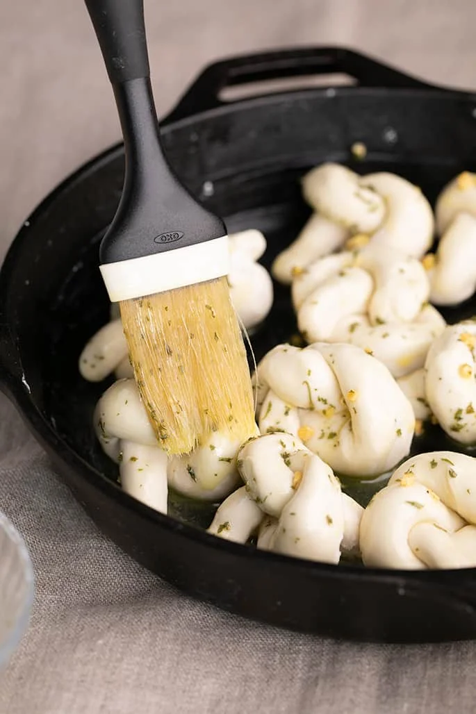 Pastry brush adding garlic and herb butter on raw shaped garlic knots in black skillet