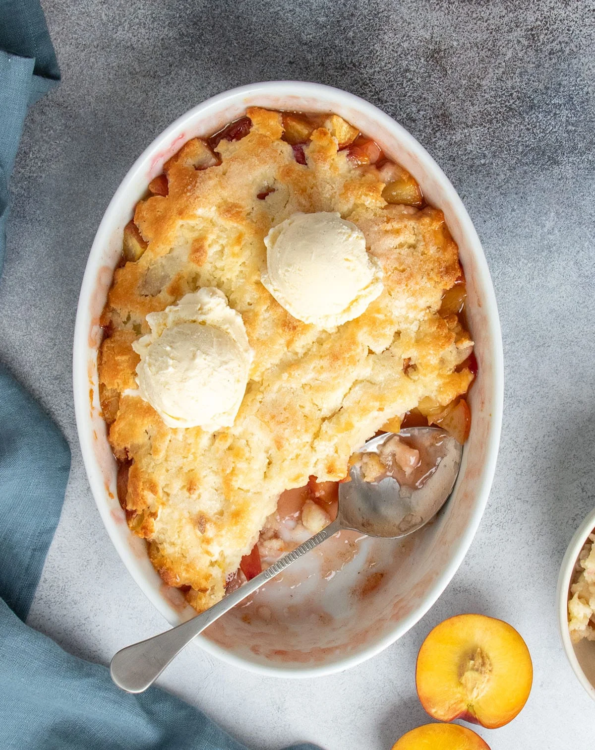 Peach cobbler in oval white serving dish served warm with scoops of vanilla ice cream melting into the golden topping.