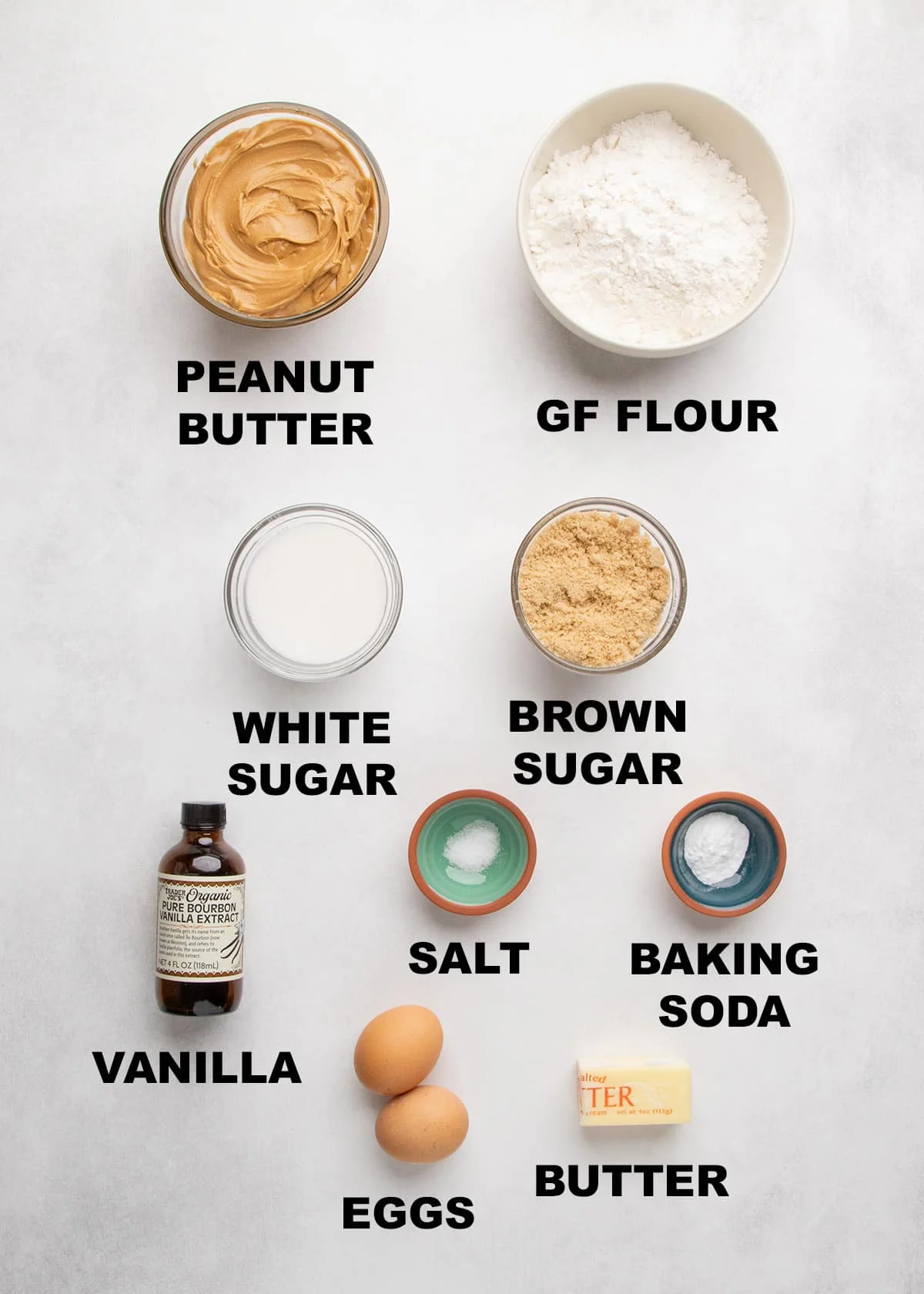 Peanut butter, gluten free flour, white sugar, brown sugar, vanilla, salt, baking soda, eggs, and butter arranged in bowls with labeled text on a light surface.
