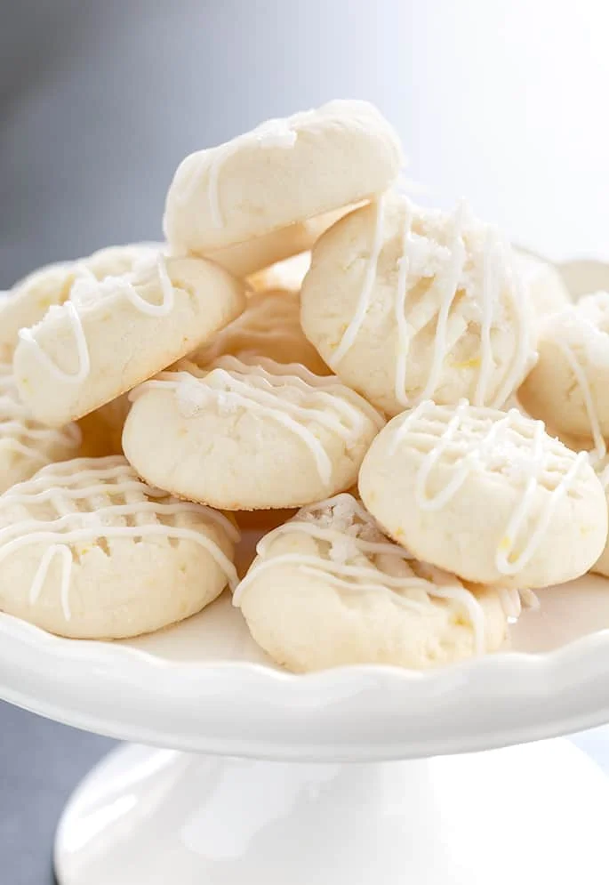Pile of small round pale cookies with white icing drizzle on small white cake plate