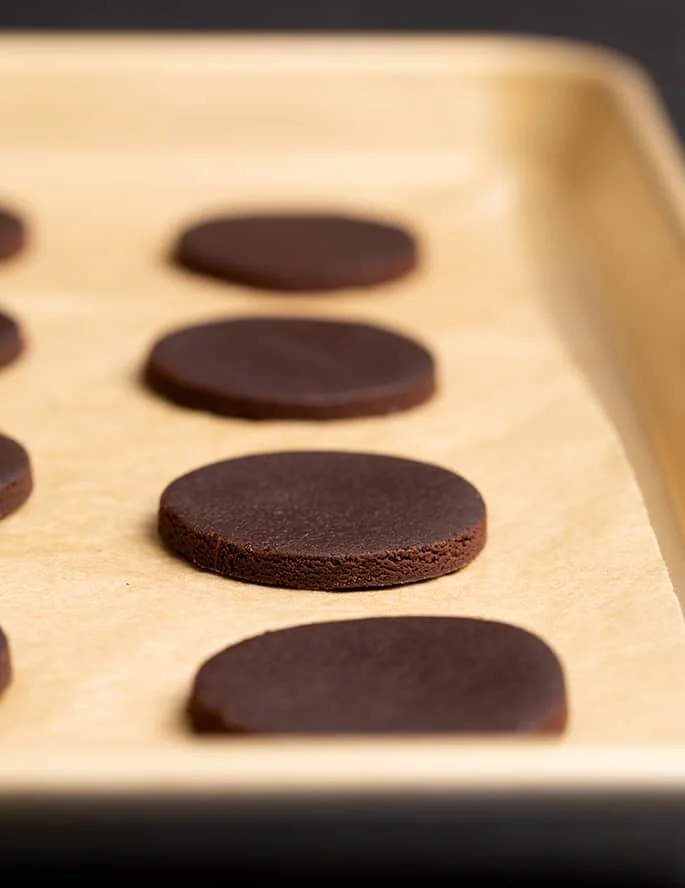 Raw brown circles of dough on brown paper on a gold colored baking tray