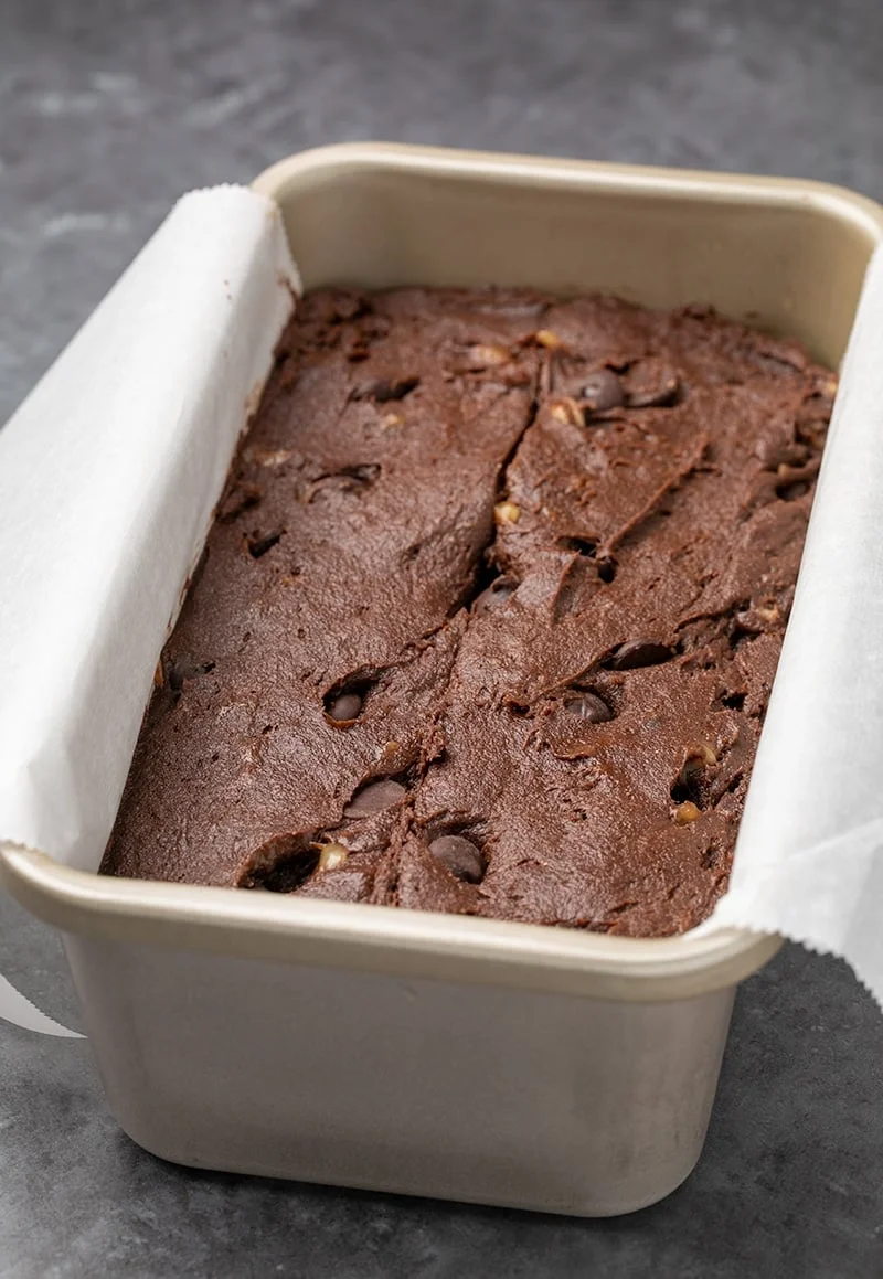 Raw dark brown batter with chips and yellow flecks in white paper in metal loaf pan.