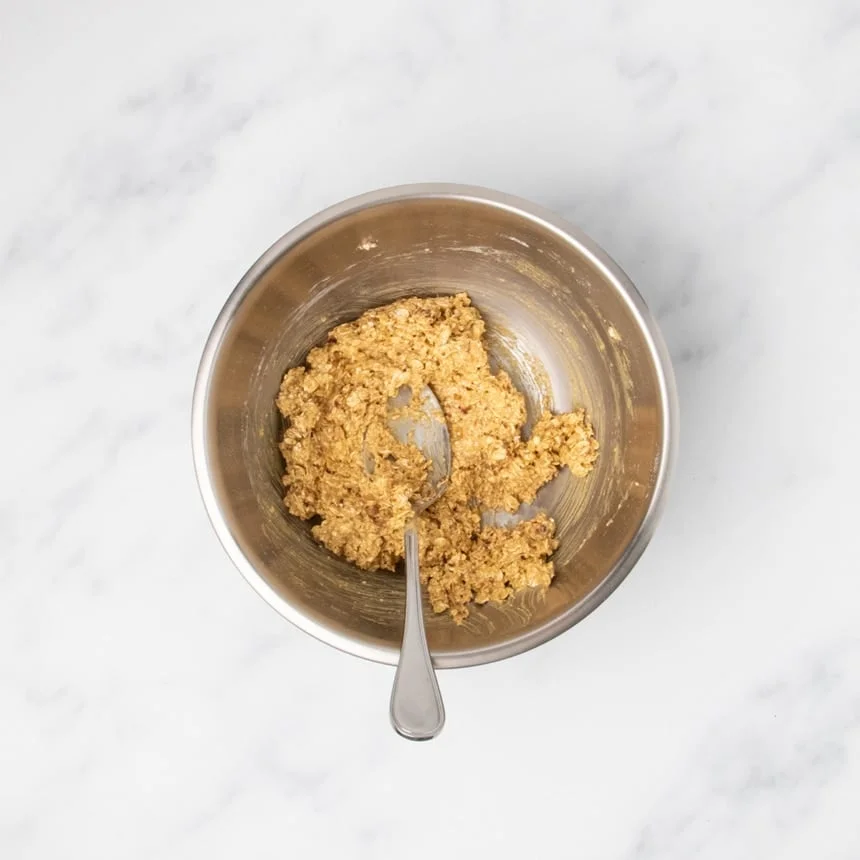 raw light brown thick raw cookie dough mixture with some rolled oats in round metal mixing bowl with large metal mixing spoon
