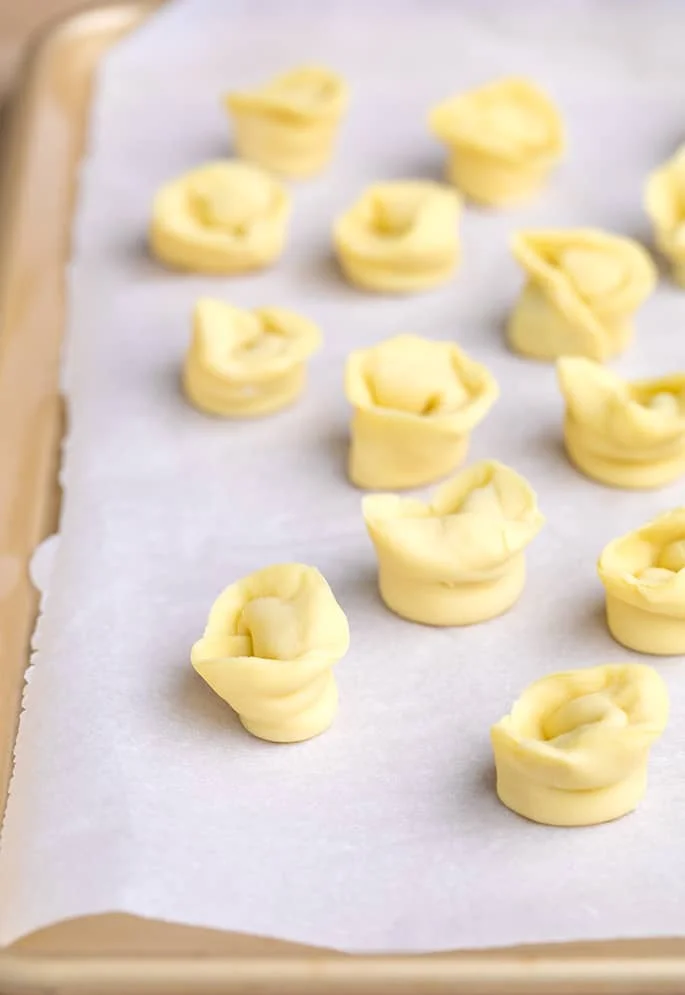 Raw shaped and filled tortellini pieces on white paper on tray.