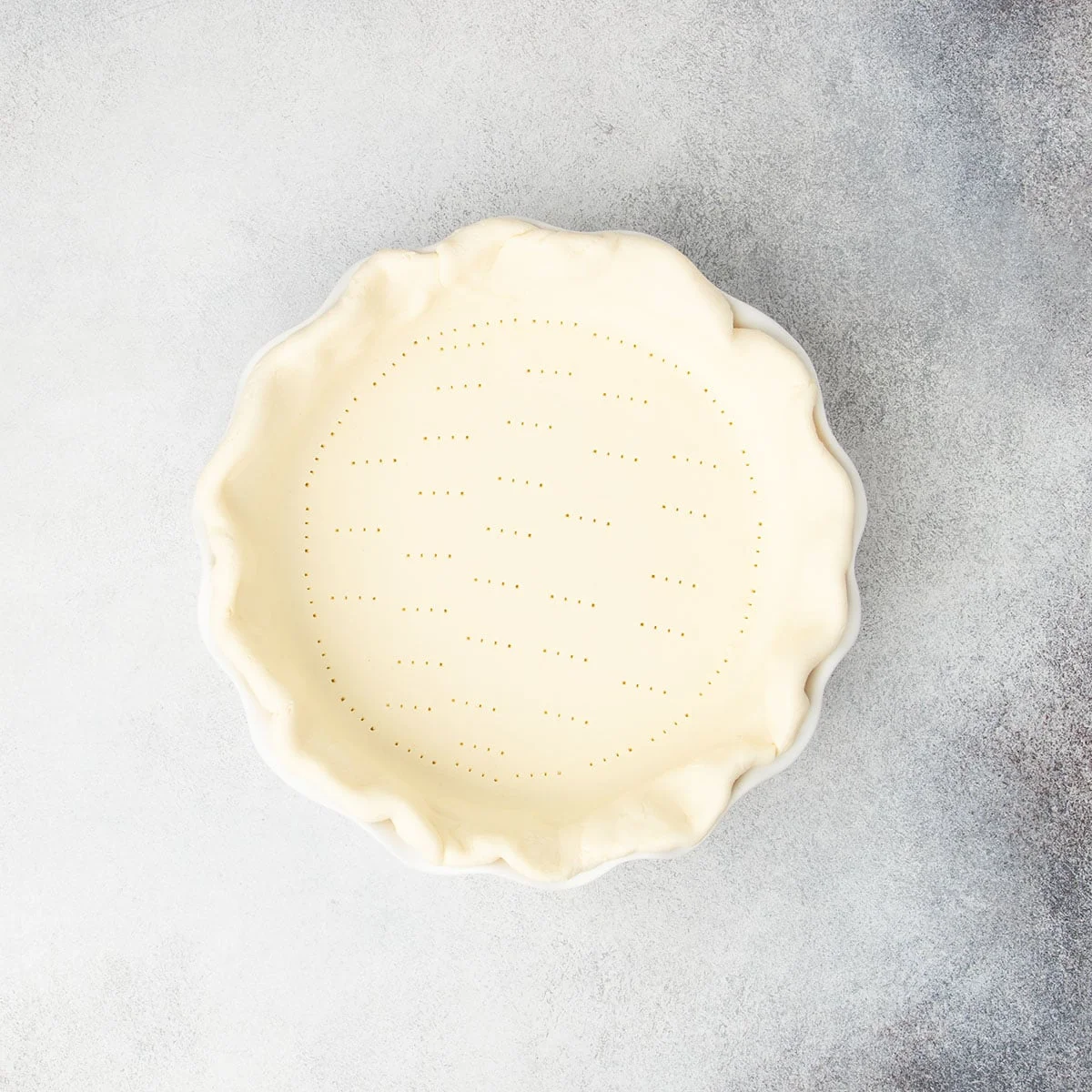 Raw shaped pie crust in white pie dish with piercings from tines of fork all over bottom of crust.