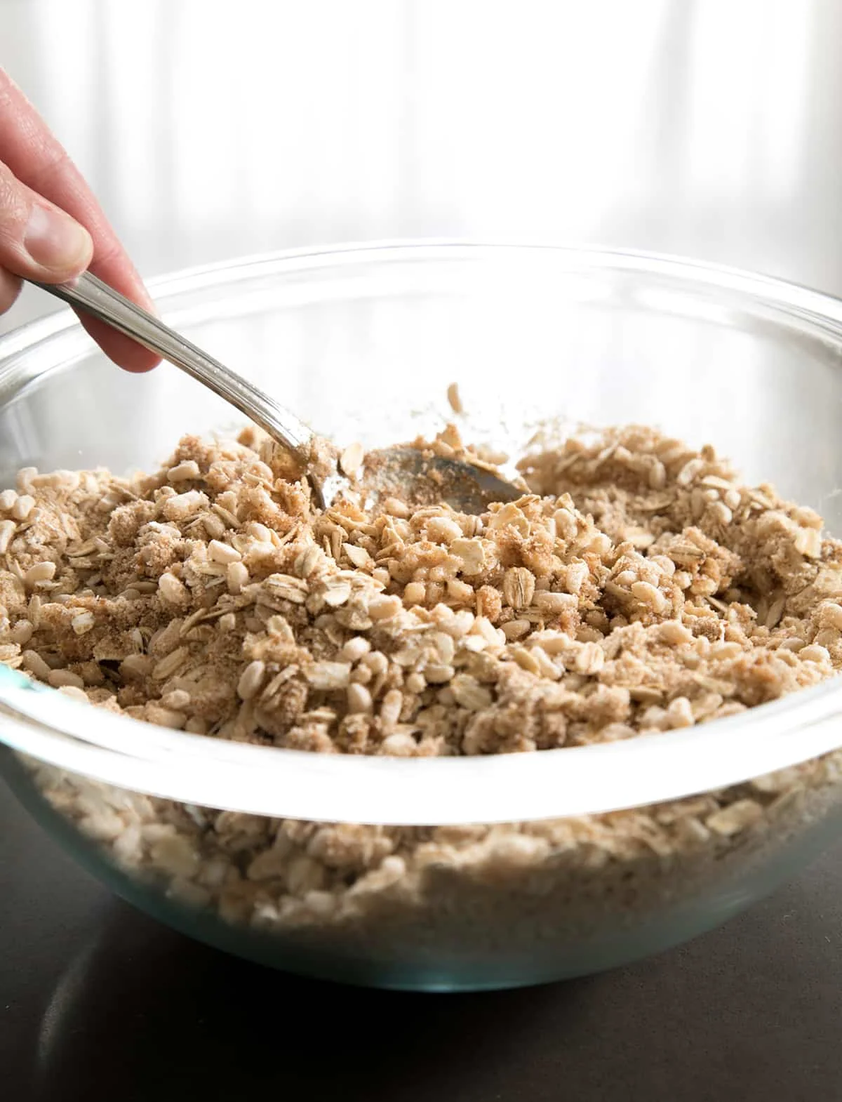 raw unshaped mixture for crunchy granola bars in glass mixing bowl with hand holding mixing spoon