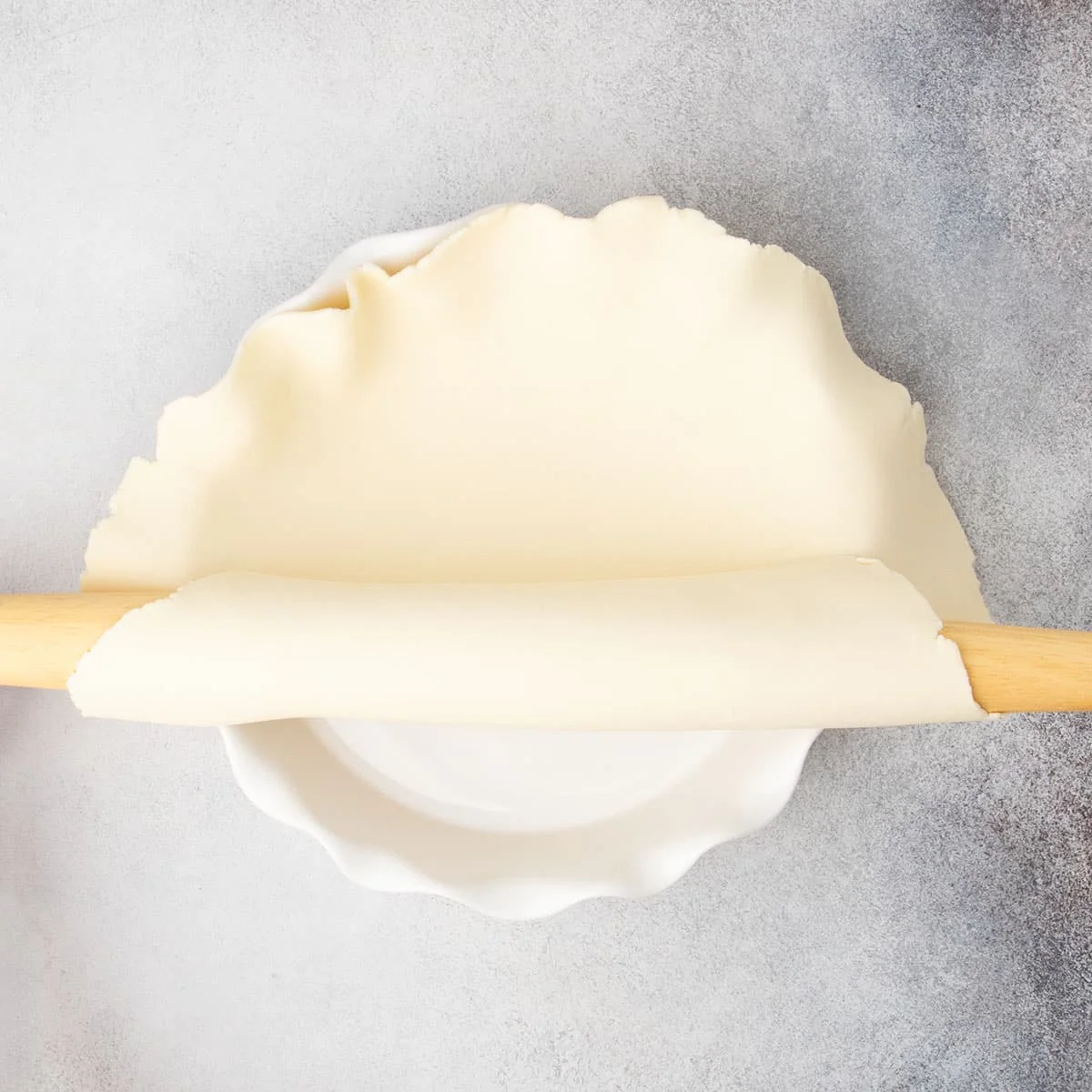 Rolled round of raw pie crust being rolled off of blonde wood rolling pin onto white porcelain scalloped edge pie dish.