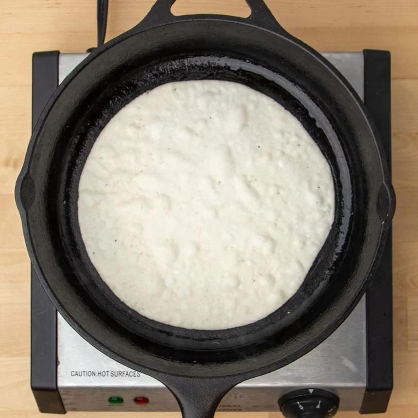 round black skillet on electric burner cooking underside of otherwise raw round tortilla with bubbles beginnin to form through to the top
