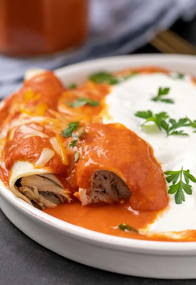 Round high sided white plate with shredded chicken inside rolled corn tortillas with enchilada sauce and parsley leaves on top with sour cream in the center