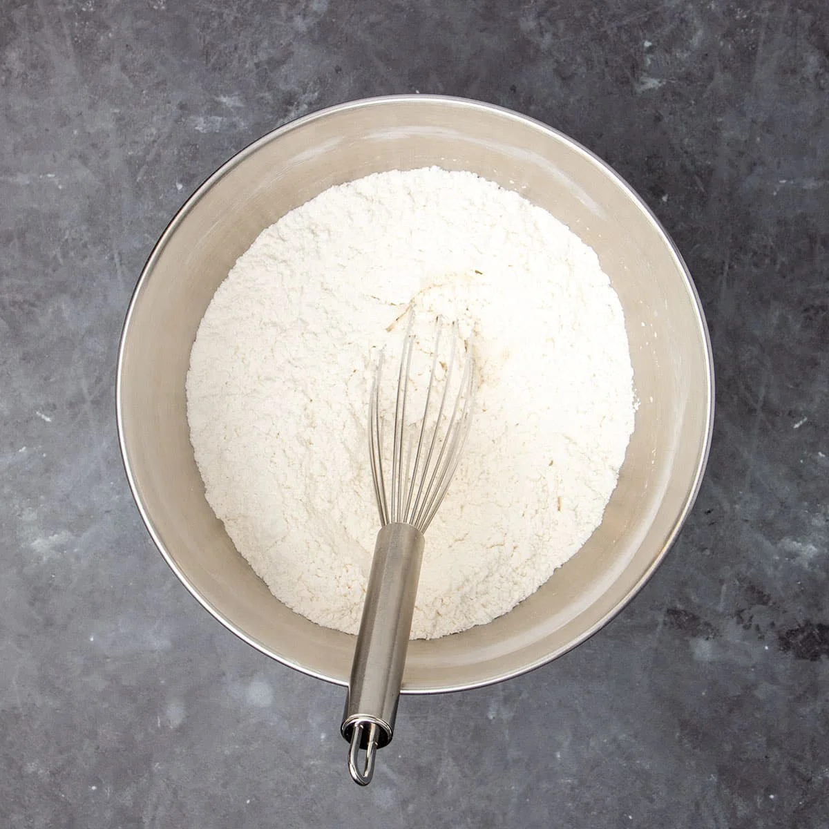 Round metal mixer bowl with white flour and metal wire whisk inside the bowl.