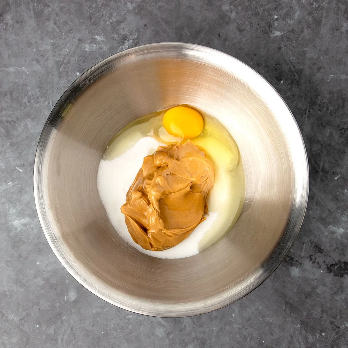 Round metal mixing bowl with light brown peanut butter, white sugar, and egg out of shell
