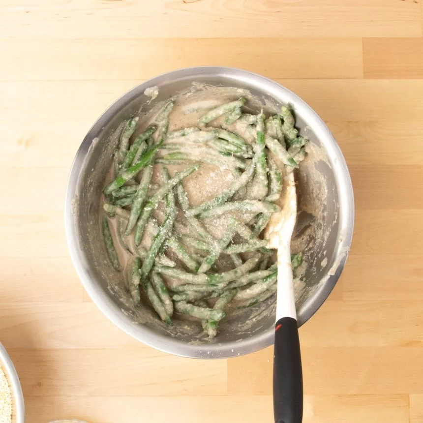 round metal mixing bowl with light brown sauce on green beans with sprinkled white grated cheese with a white a black spatula