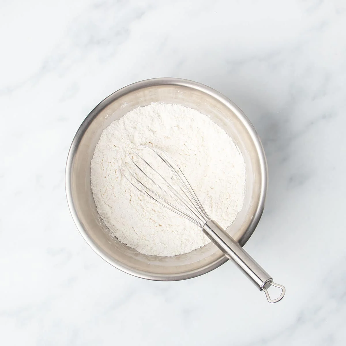 Round metal mixing bowl with white flour and wire whisk