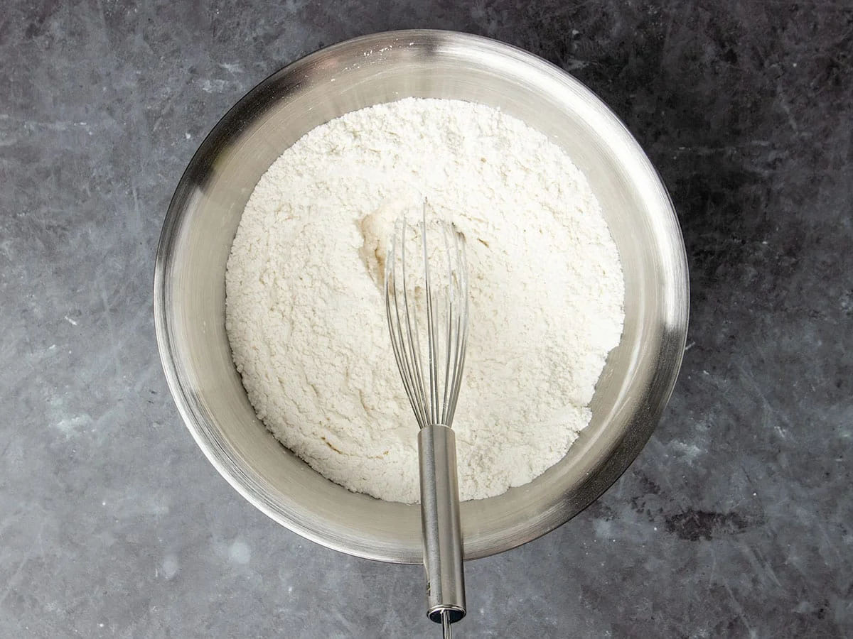 round metal mixing bowl with white flour mixture and large metal wire whisk