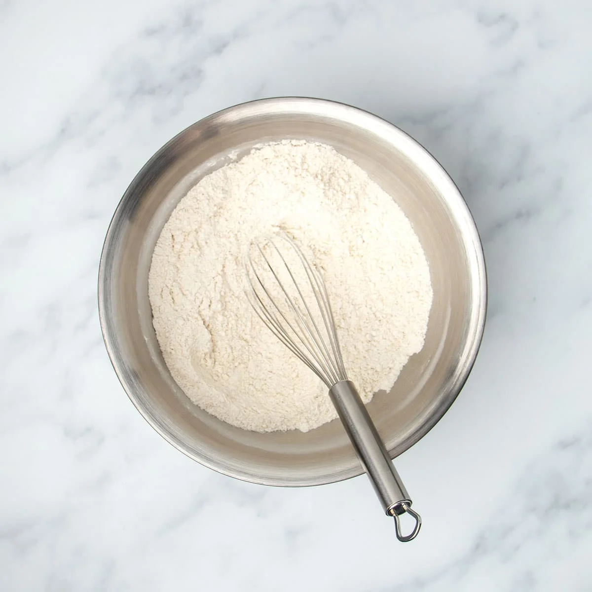 Round metal mixing bowl with white flour mixture and metal wire whisk.