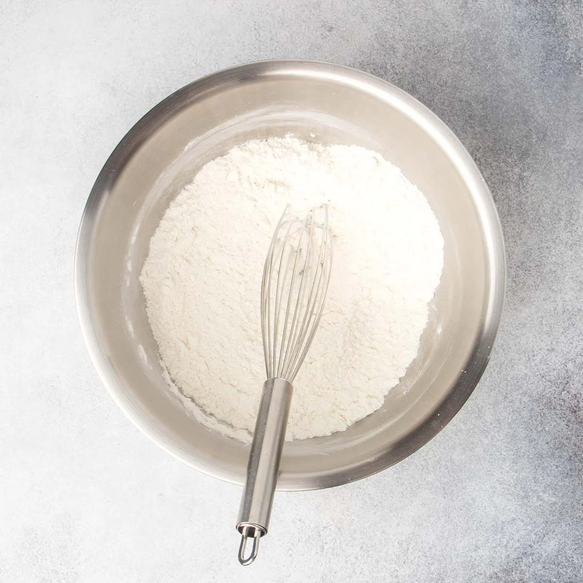 Round metal mixing bowl with white flour mixture and metal wire whisk.