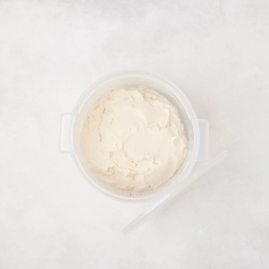 Round plastic container filled with raw smooth pale dough and a lid placed beside it on a light surface.