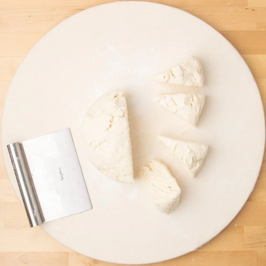 Round white board with a large piece of disk-shaped dough cut in half and several triangular pieces arranged nearby, with a metal bench scraper on the side.