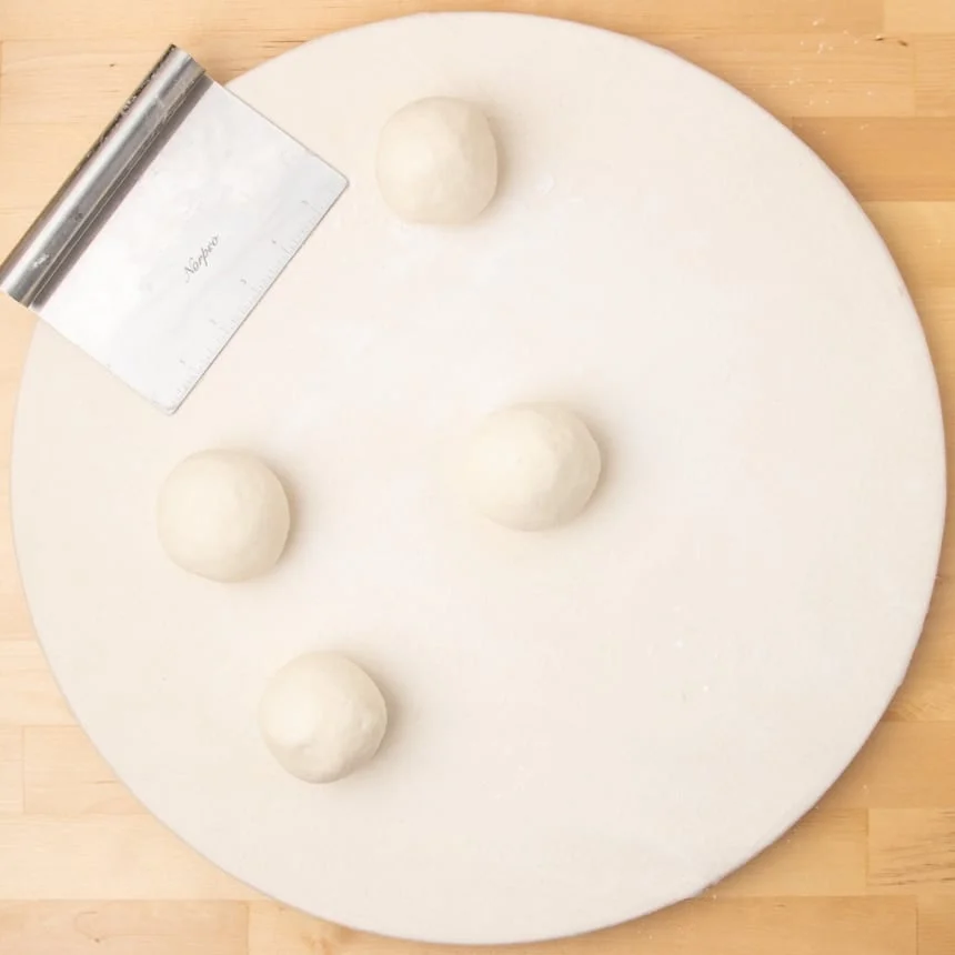 Round white board with four smooth dough balls spaced apart and a metal bench scraper resting nearby.