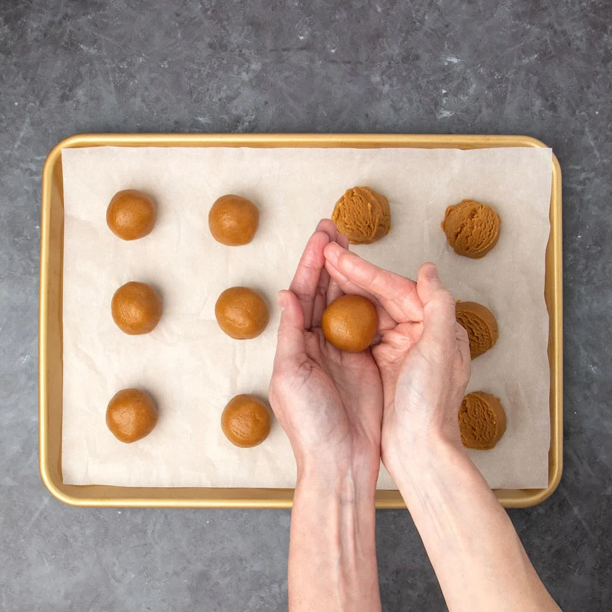 same baking sheet with about half the mounds of cookie dough rolled into rounds with one round piece of cookie dough in the palms of 2 hands