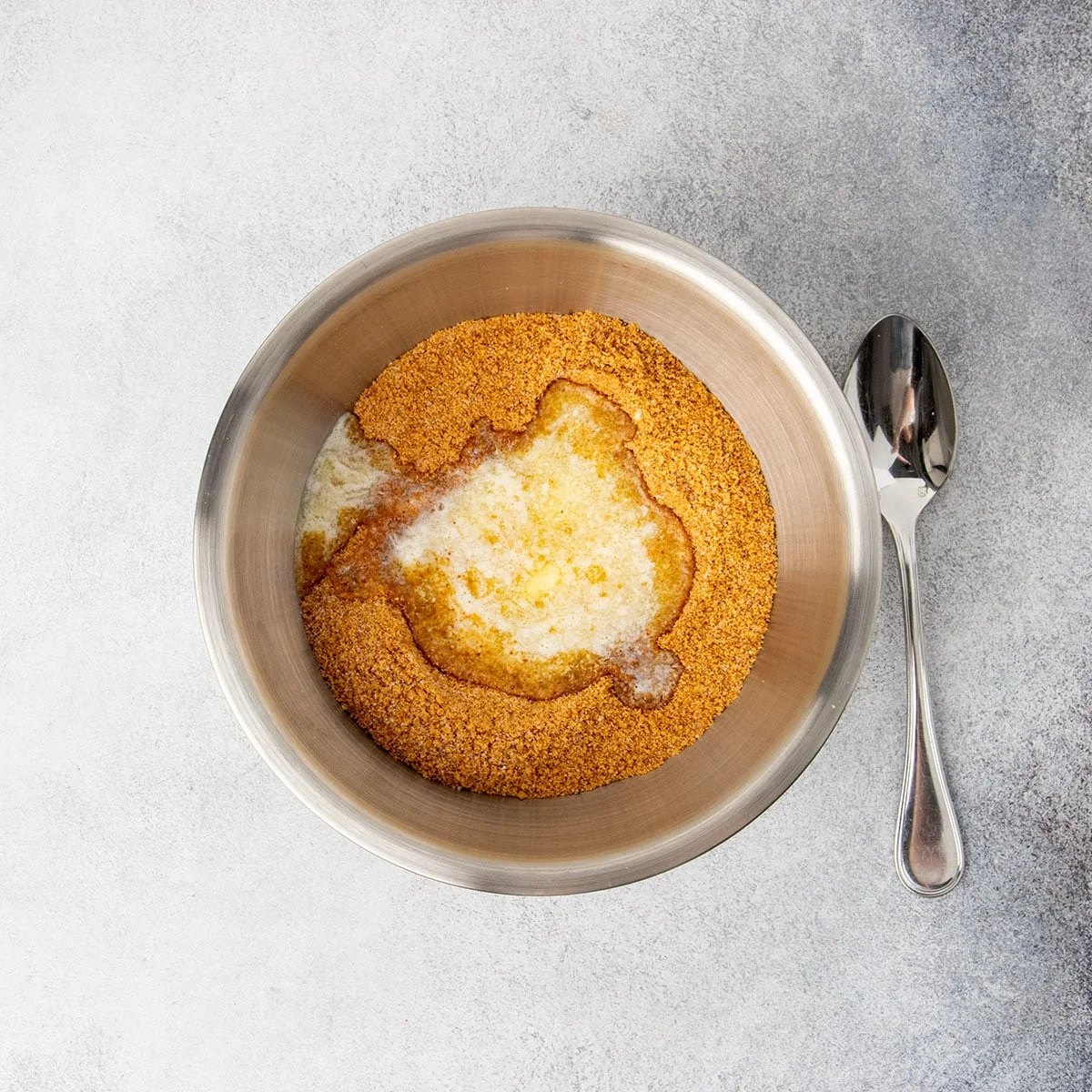Same bowl with brown crumbs whisked with white sugar and melted yellow butter sitting on top with a spoon next to the bowl.