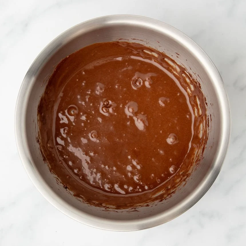 same bowl with darker brown mixture of Nutella and eggs mixed together, with some bubbles on top of mixture
