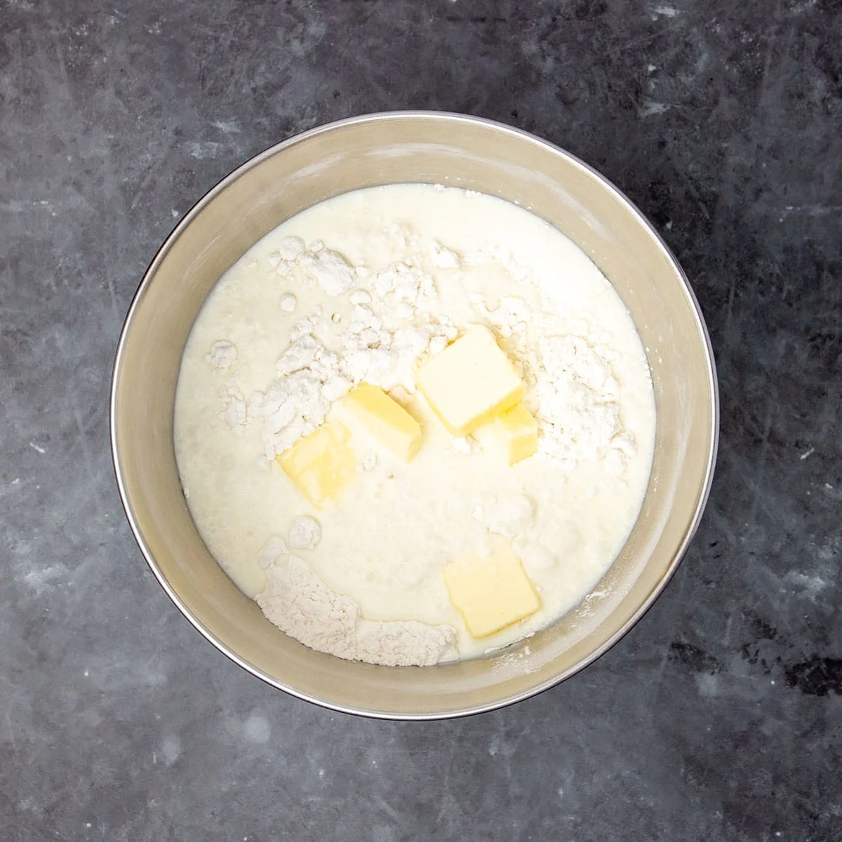 Same bowl with yellow pats of butter, white milk and light brown liquid sourdough starter on top of dry flour.