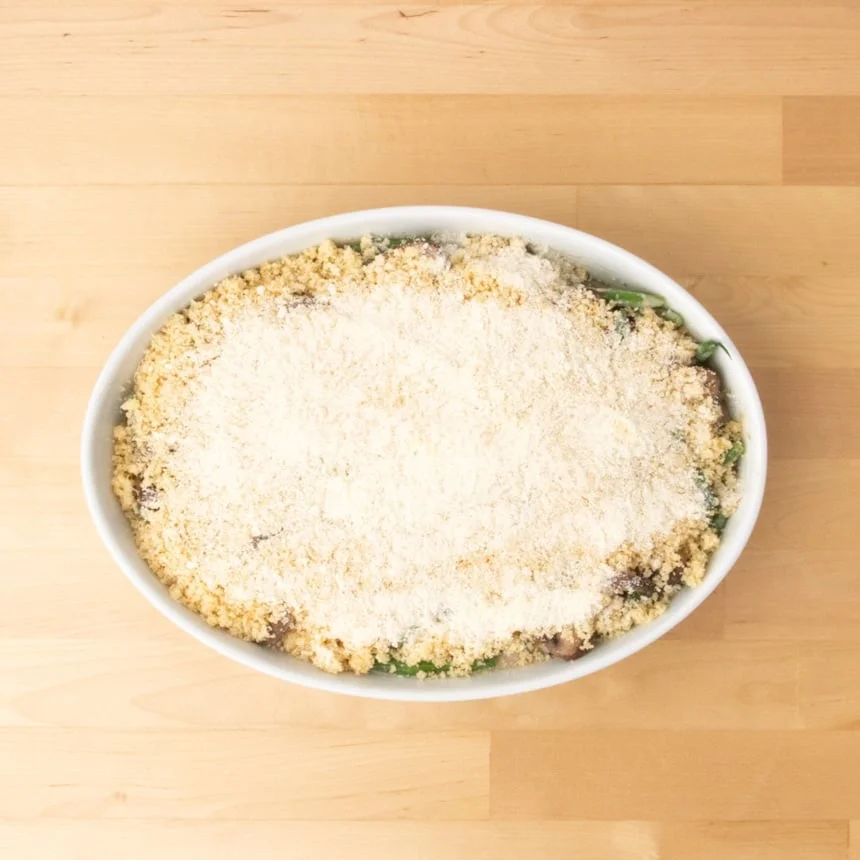 same casserole dish with the prepared breadcrumb mixture and a top layer of white grated cheese, ready to go in the oven