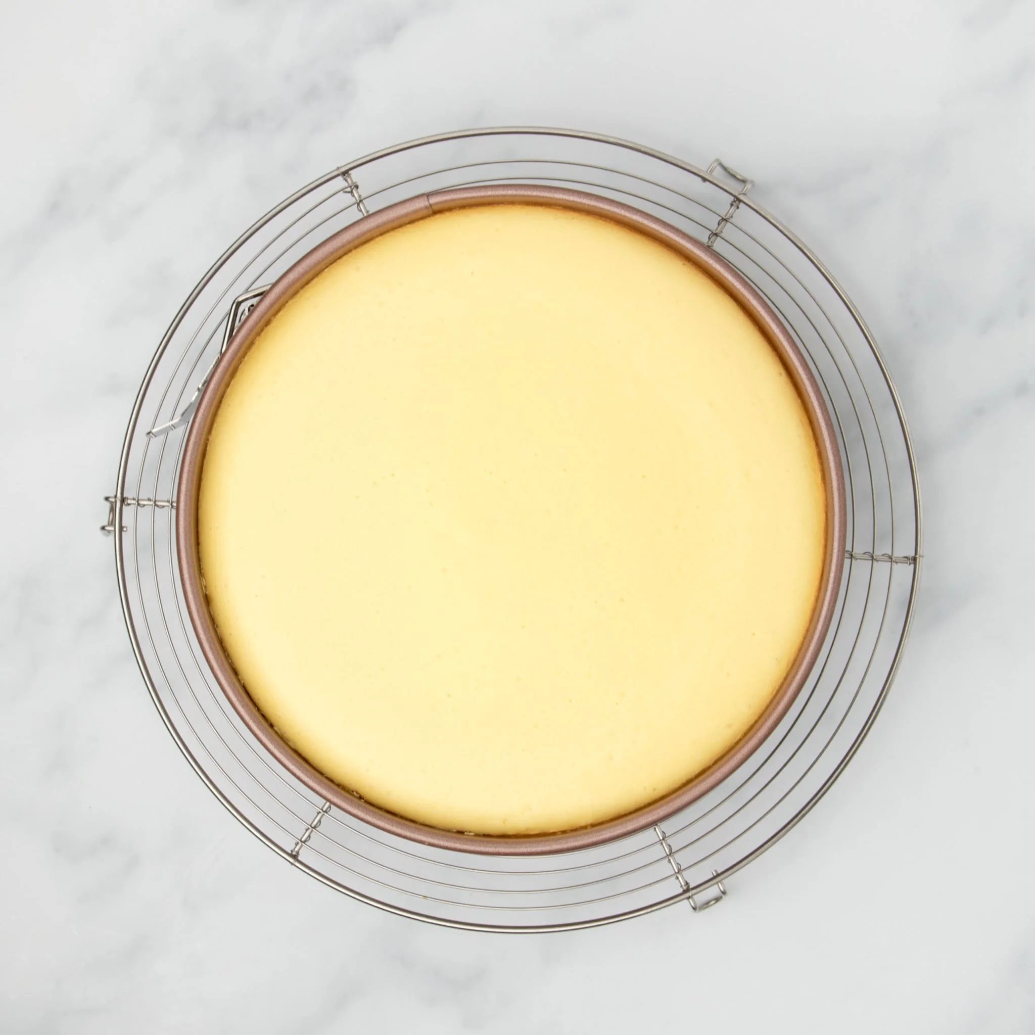 same cheesecake mixture in gold springform pan now baked into slightly darker yellow cake on round wire rack