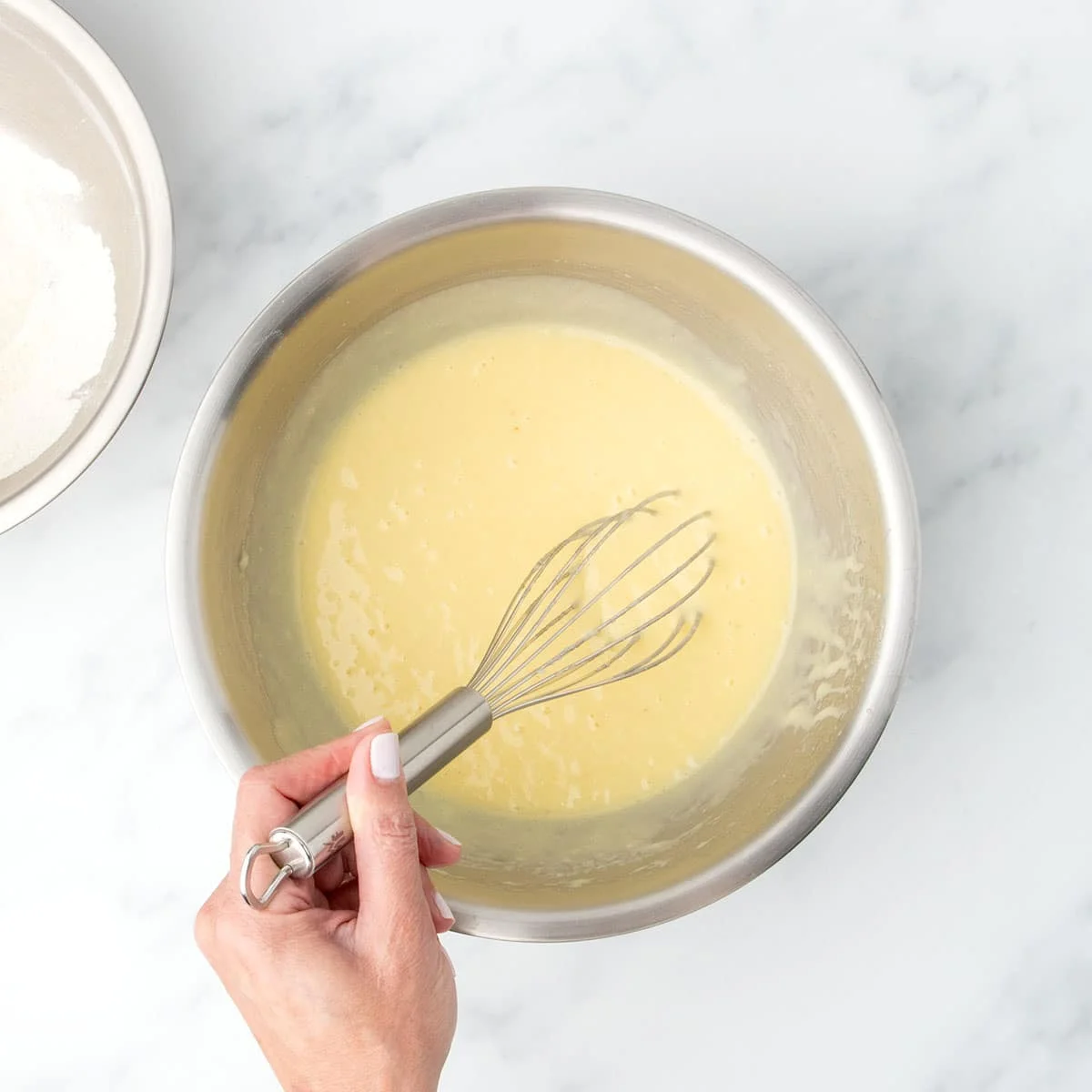 Same metal mixing bowl with some dry ingredients and all wet ingredients forming a pale yellow batter, with a whisk held in hand.