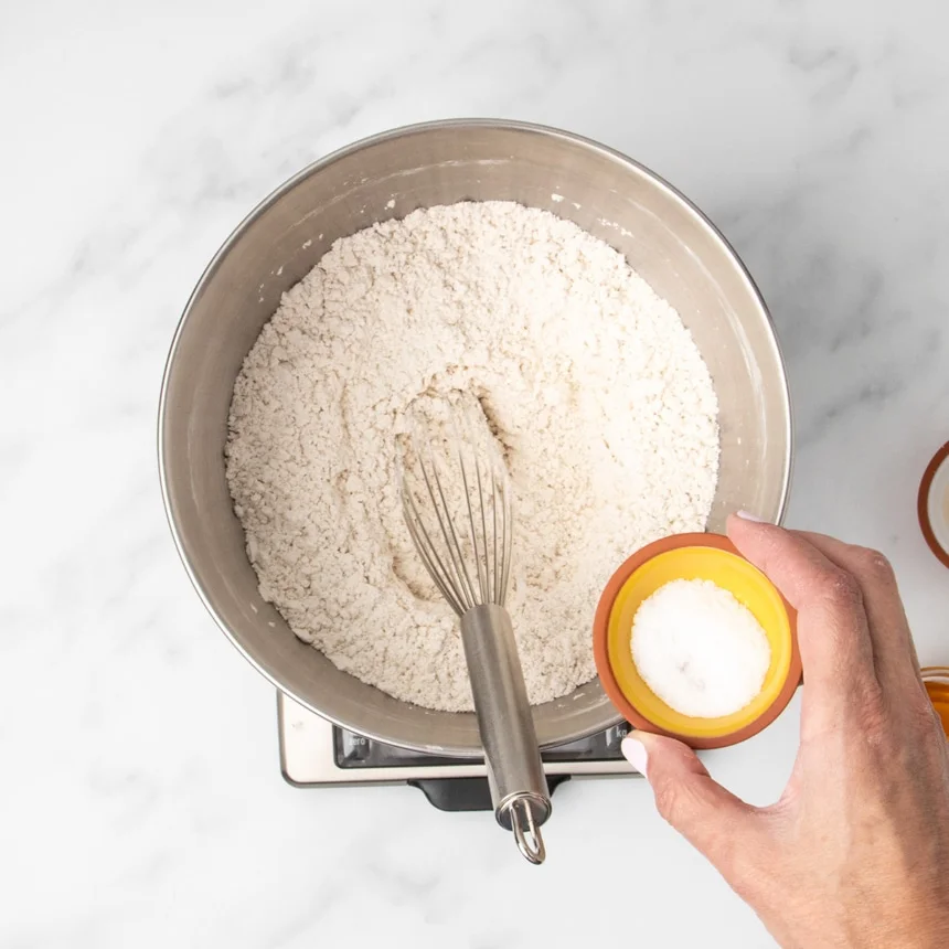 same mixing bowl, now with hand holding pinch bowl with coarse salt about to be added, with wire whisk in bowl