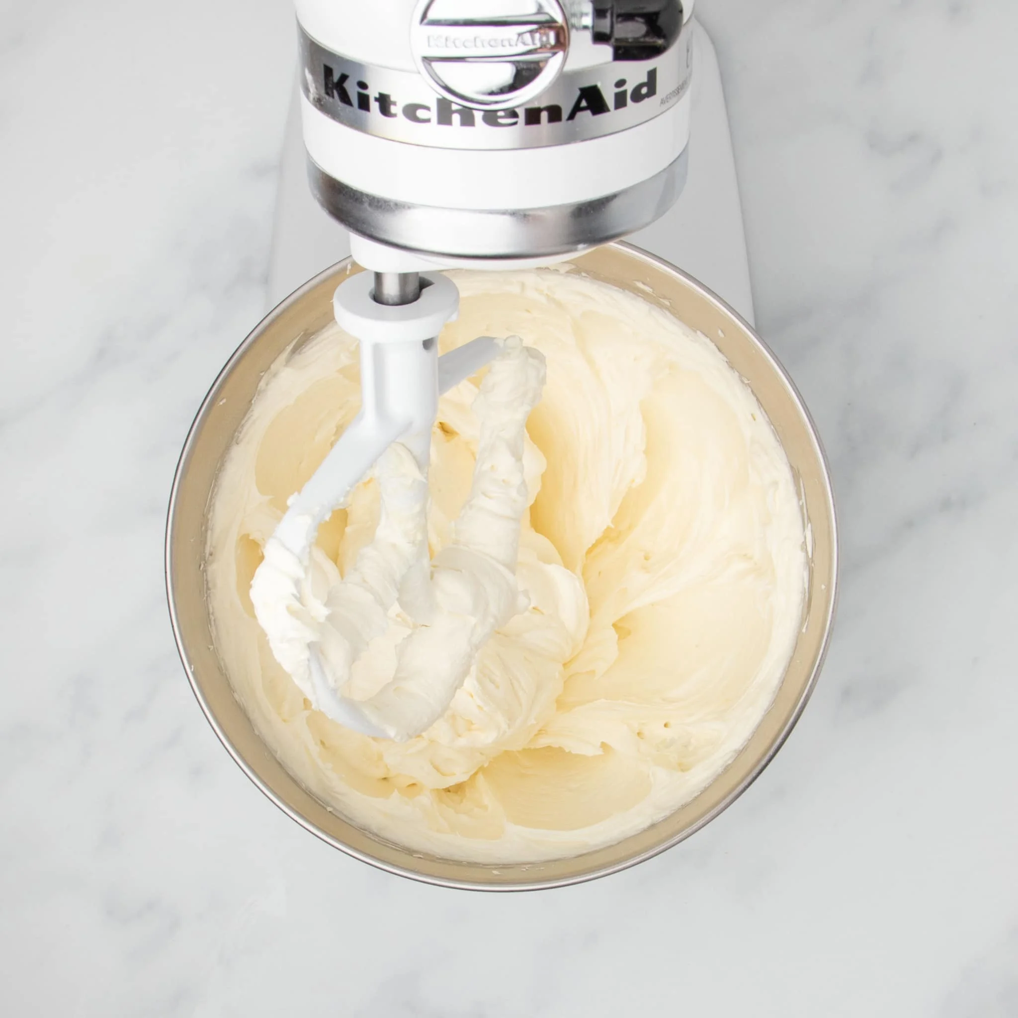 same mixing bowl with cream colored fluffy mixture of beaten cream cheese with white paddle attachment and white kitchenaid mixer