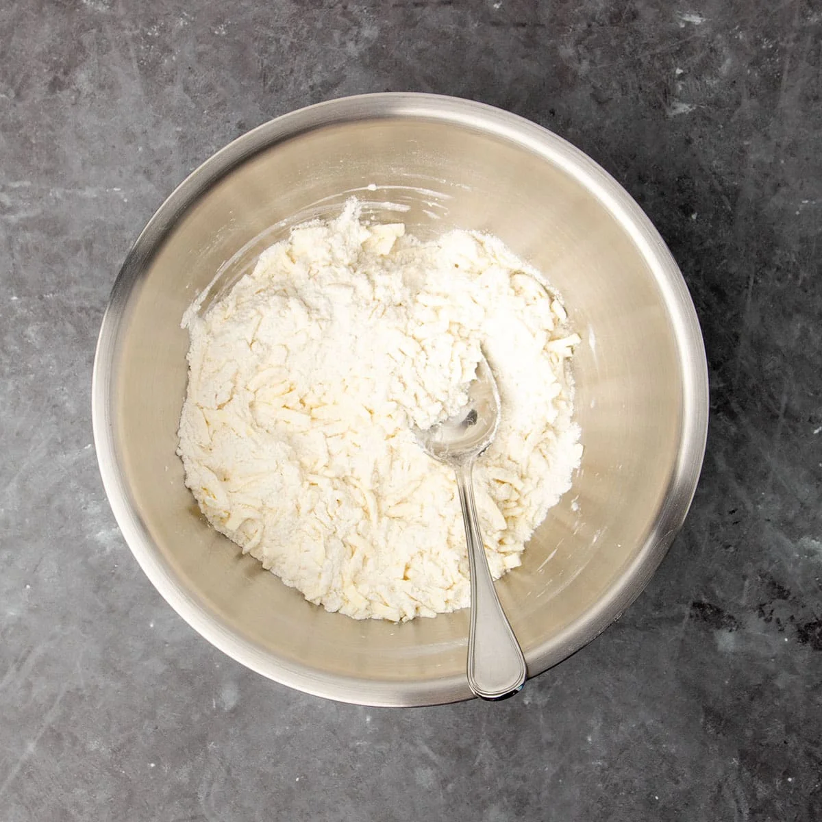 same mixing bowl with grated light yellow butter shreds mixed in to flour with large metal mixing spoon