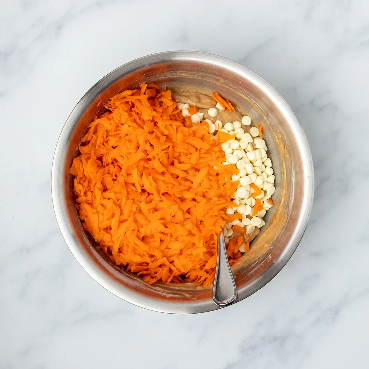 Same mixing bowl with light brown batter underneath and shredded orange carrots and white chocolate chips on top with metal mixing spoon.