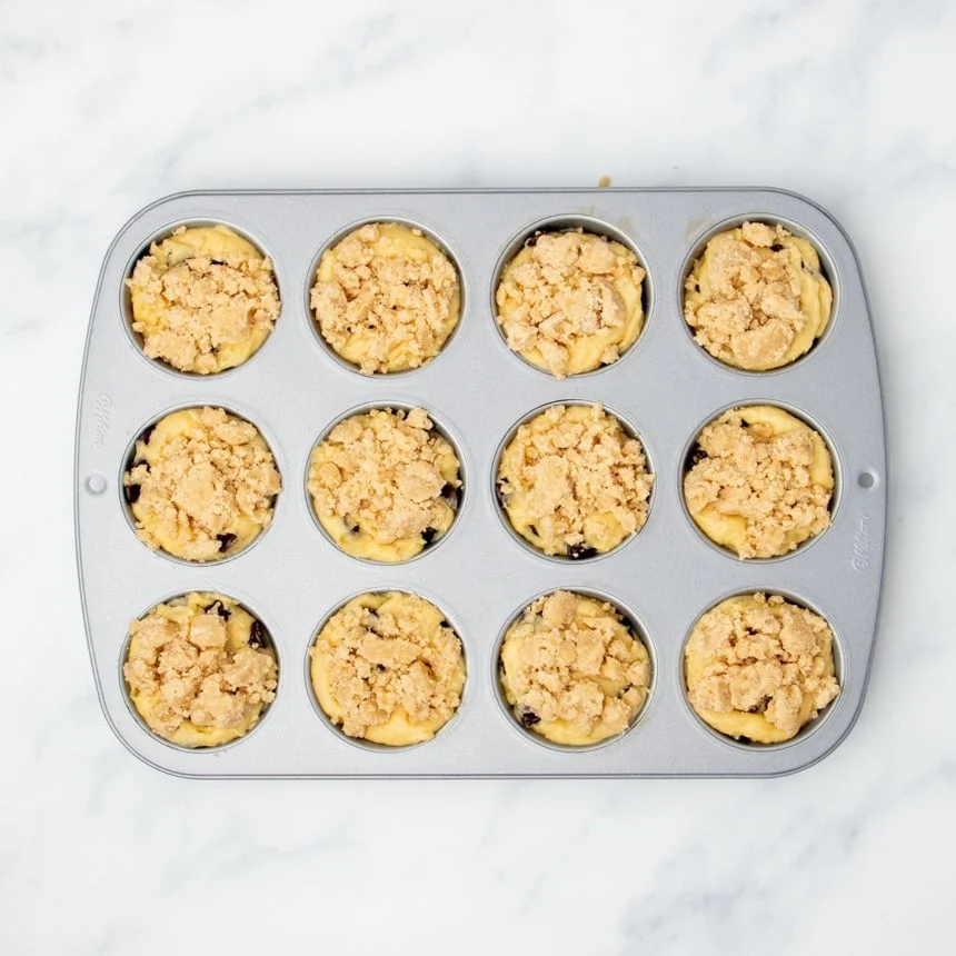 same muffin tin with light brown clumps of chilled crumble mixture on top of batter in each well
