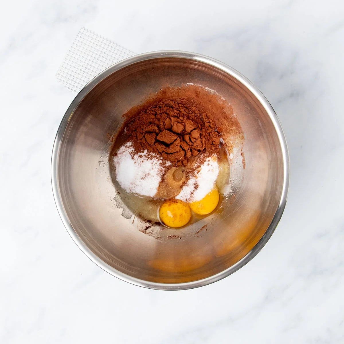 Separate round metal mixing bowl with eggs, sugar, cocoa powder, vanilla, and salt.