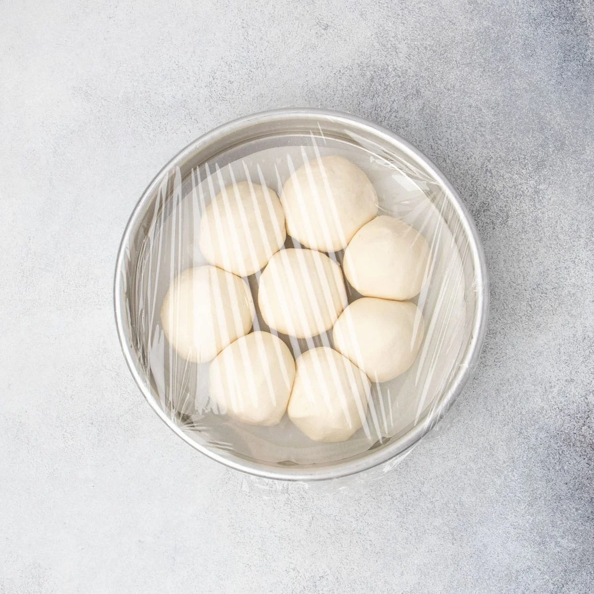 Shaped raw round rolls arranged in a round baking pan, covered with plastic wrap.