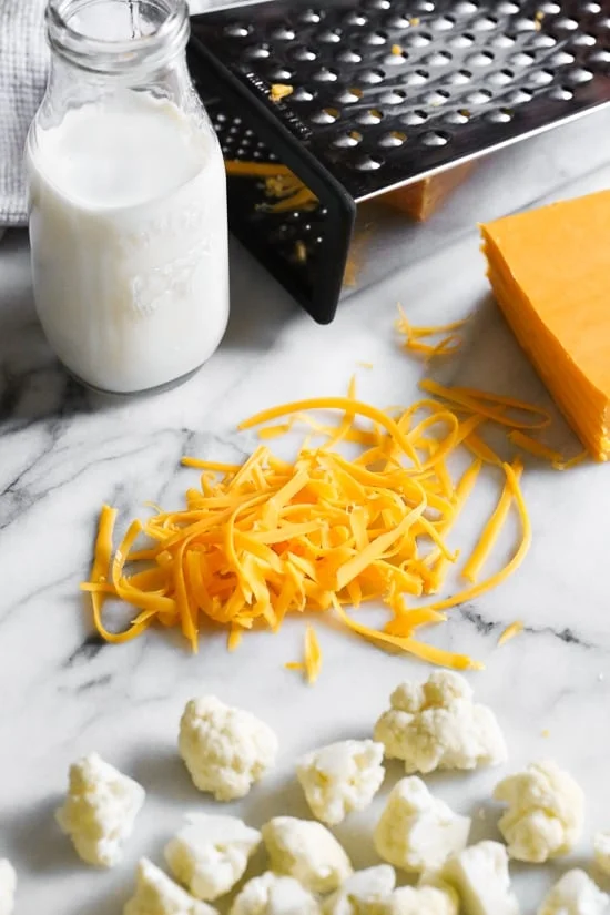 Shredded cheddar cheese on a countertop next to cauliflower florets.