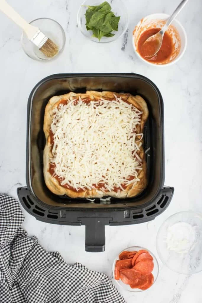 Shredded cheese layered on top of pizza dough & sauce in a black air fryer basket.