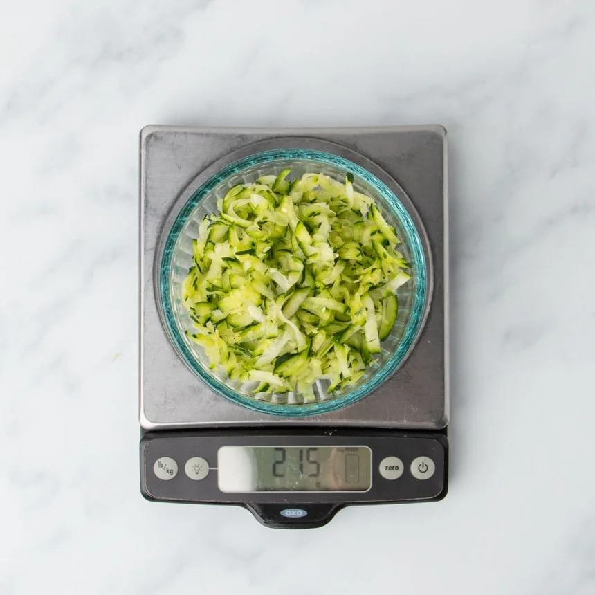 shredded zucchini in small round glass bowl on metal digital kitchen scale with 215 g in display