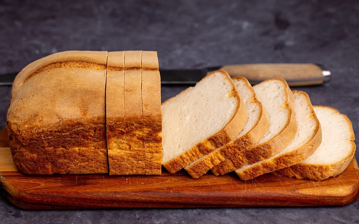 Side image of loaf of bread with brown crust and white crumb with 8 slices cut, and whole thing sitting on a brown bread board, with a bread knife in background.