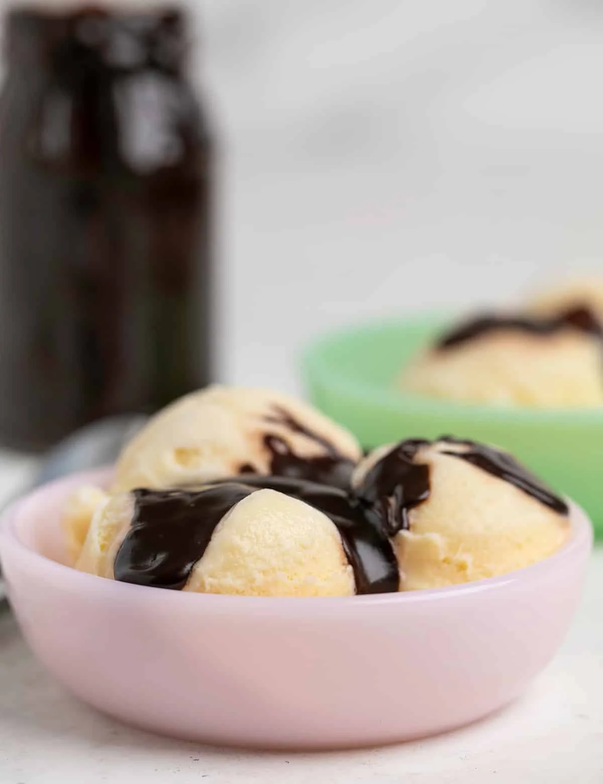 Side view of a small pink bowl with 3 scoops of vanilla ice cream with hot fudge sauce on top and in a jar in the background