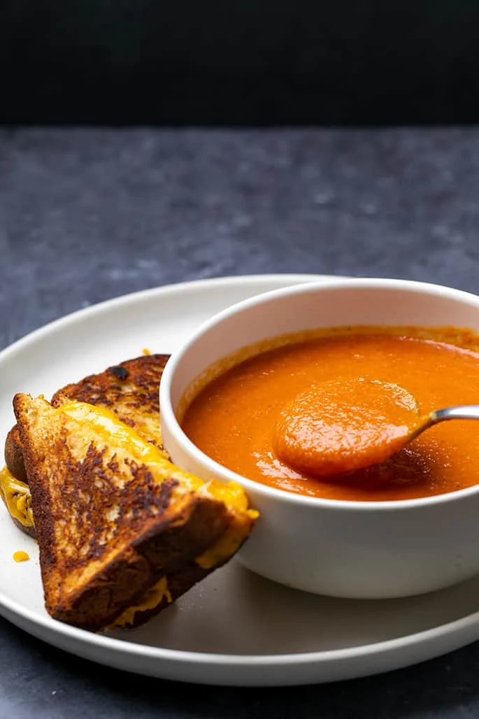 Side view of spoon above bowl of tomato soup with grilled cheese sandwich on side