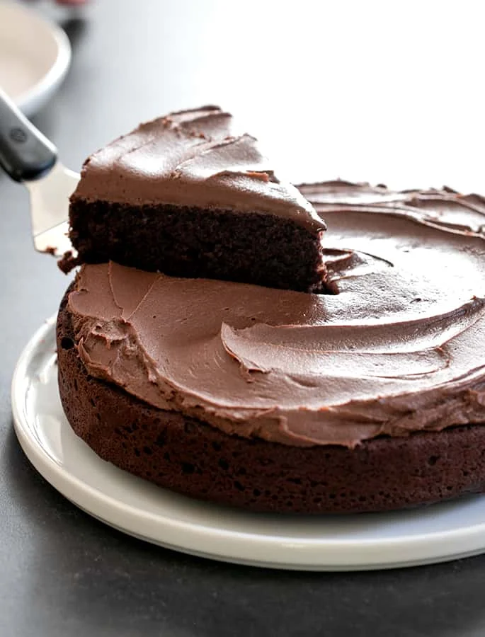 single layer brown gluten free chocolate cake with brown chocolate frosting on a white serving platter with a spatula lifting one slice of cake from the rest