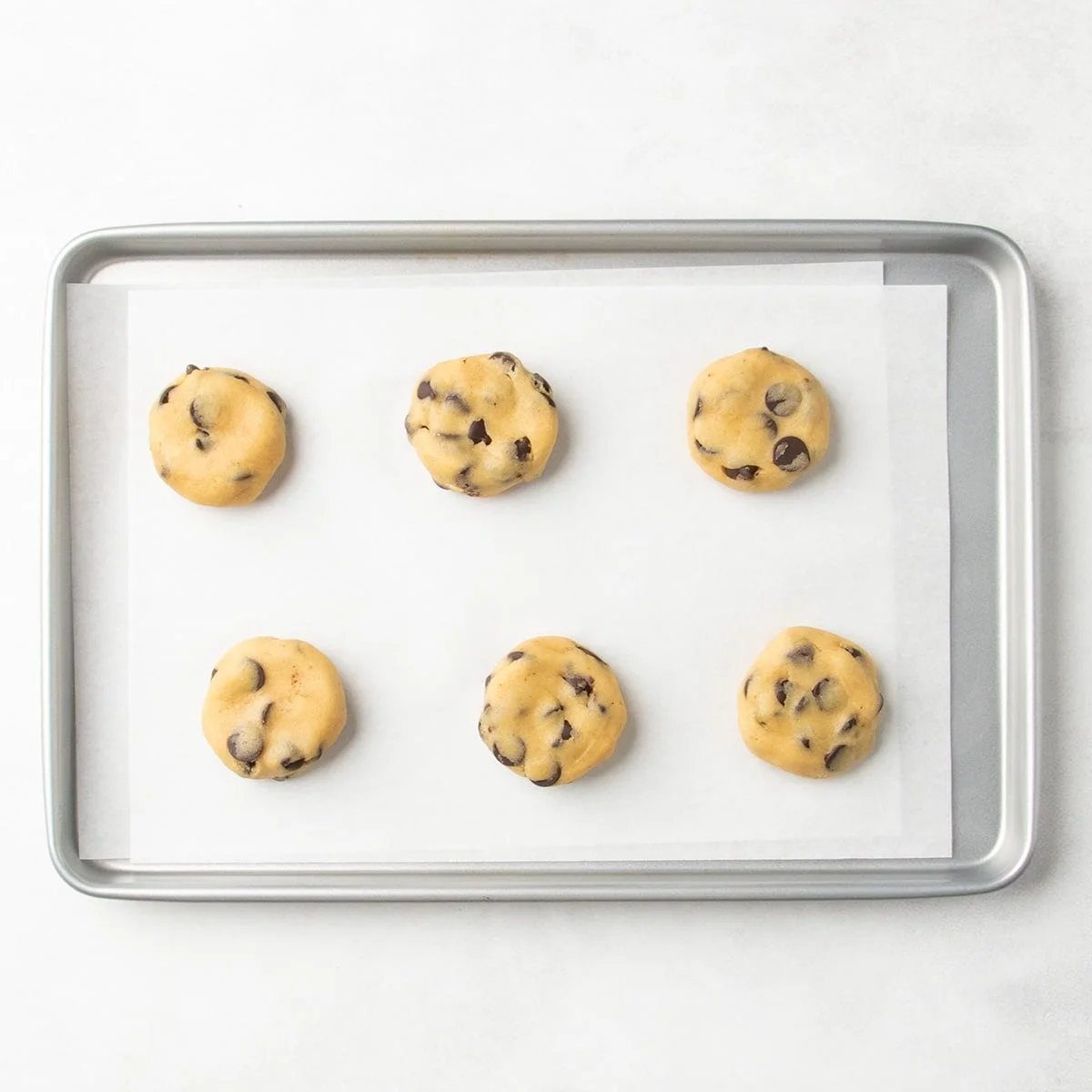 Six flattened chocolate chip cookie dough portions spaced apart on a parchment-lined baking sheet.