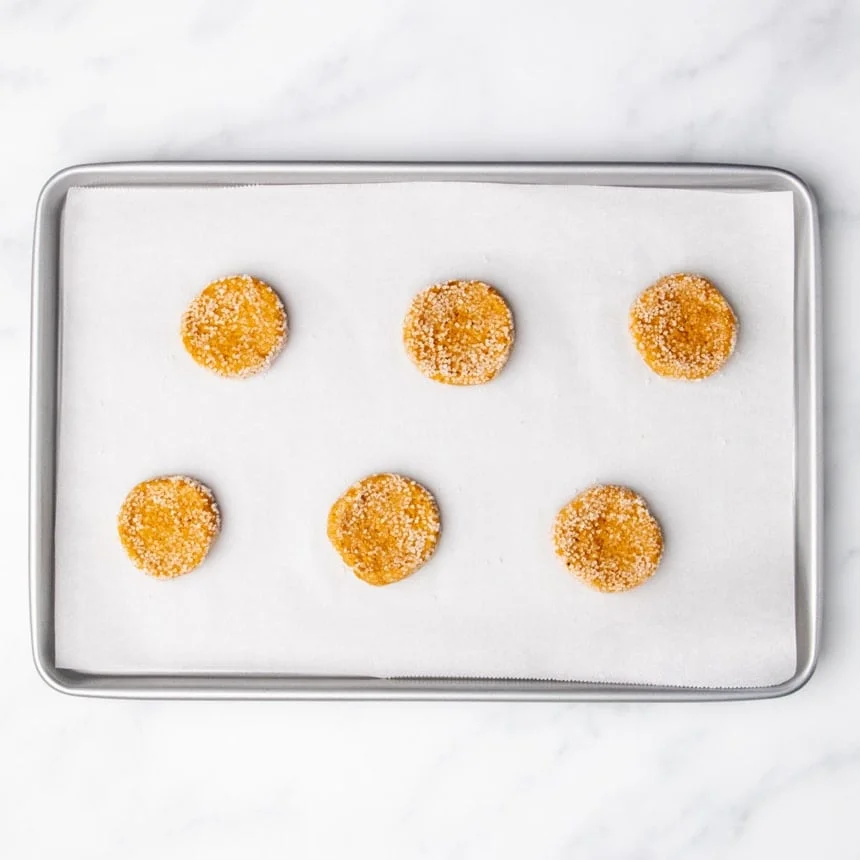 Six sugar-coated cookie dough rounds spaced apart on a parchment-lined baking sheet.